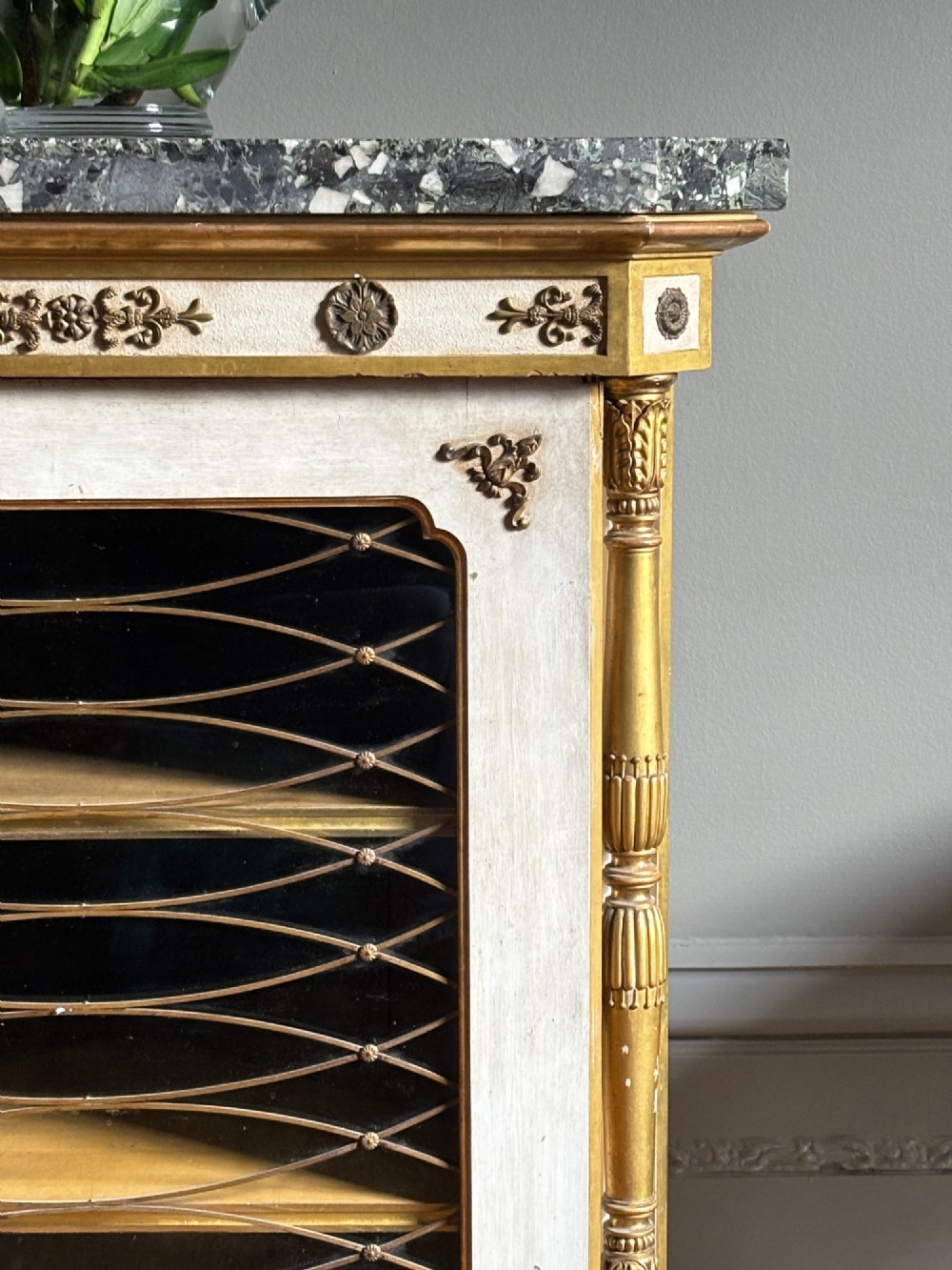 An Italian Regency period corner cupboard featuring a grey marble top, cream painted door with a gilt metal grille, fluted side columns, and decorative gilt rosettes. View III
