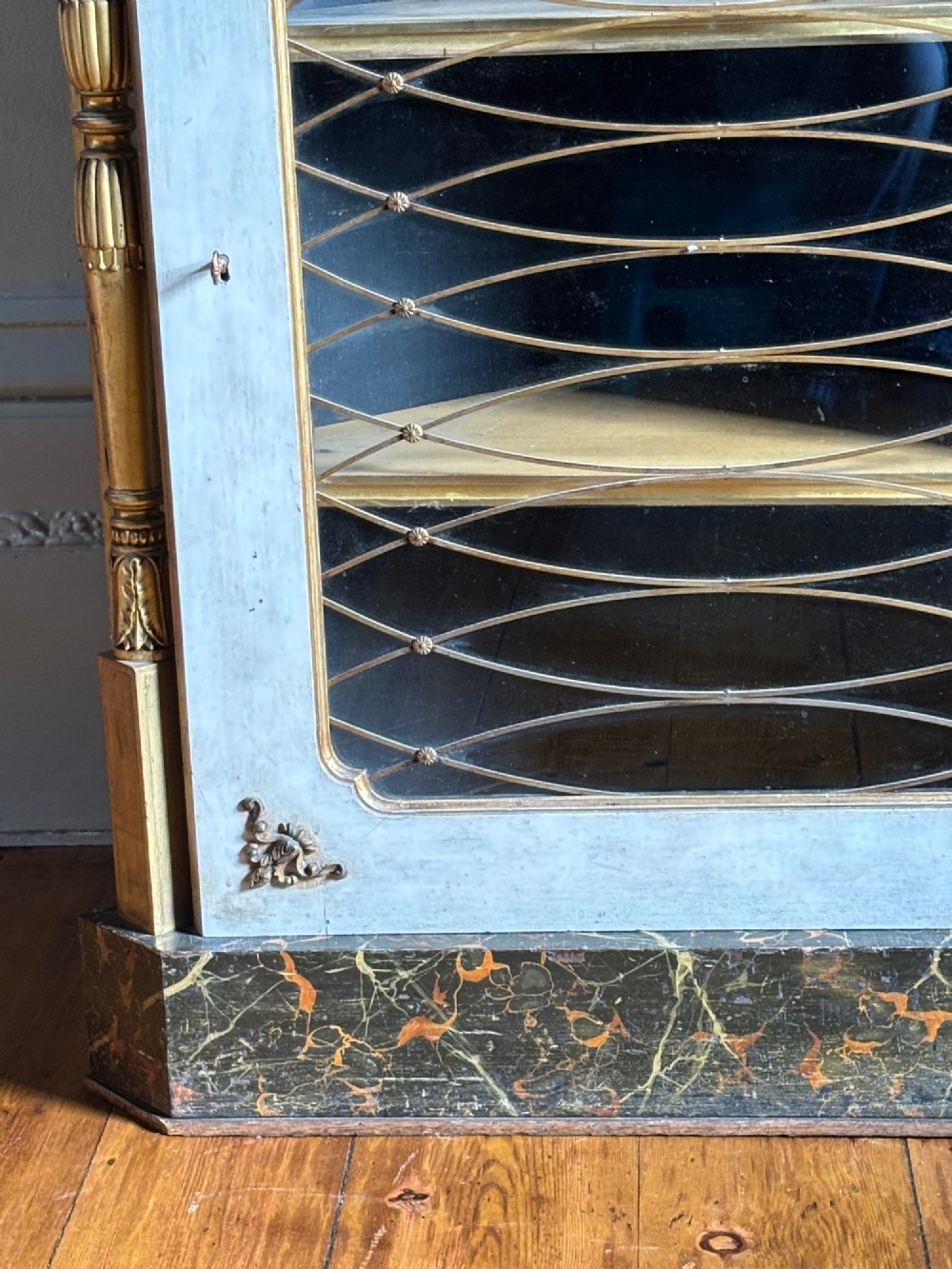 An Italian Regency period corner cupboard featuring a grey marble top, cream painted door with a gilt metal grille, fluted side columns, and decorative gilt rosettes. View IV