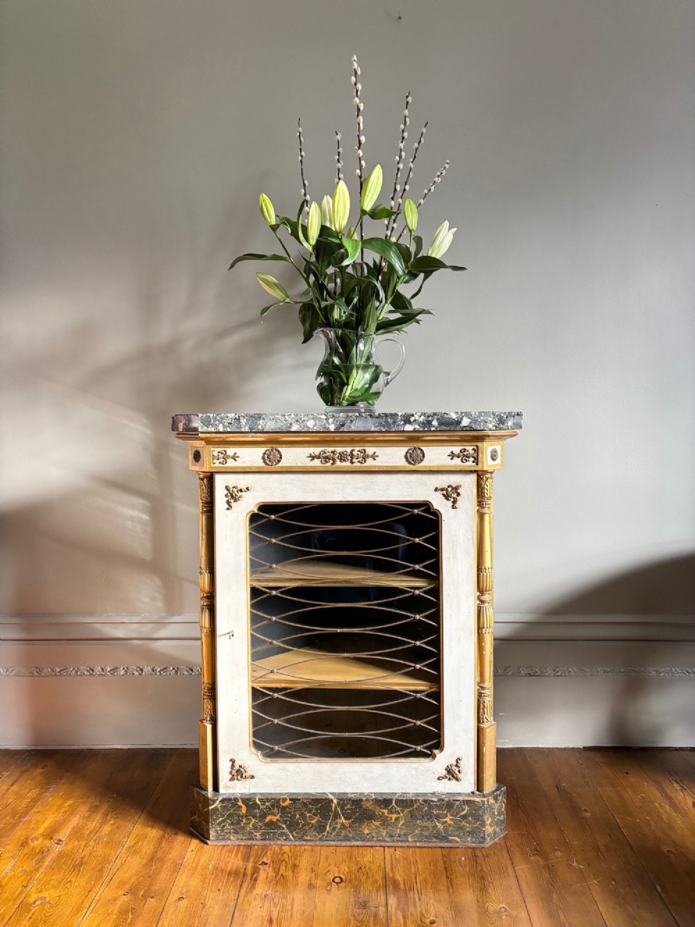 An Italian Regency period corner cupboard featuring a grey marble top, cream painted door with a gilt metal grille, fluted side columns, and decorative gilt rosettes. View V