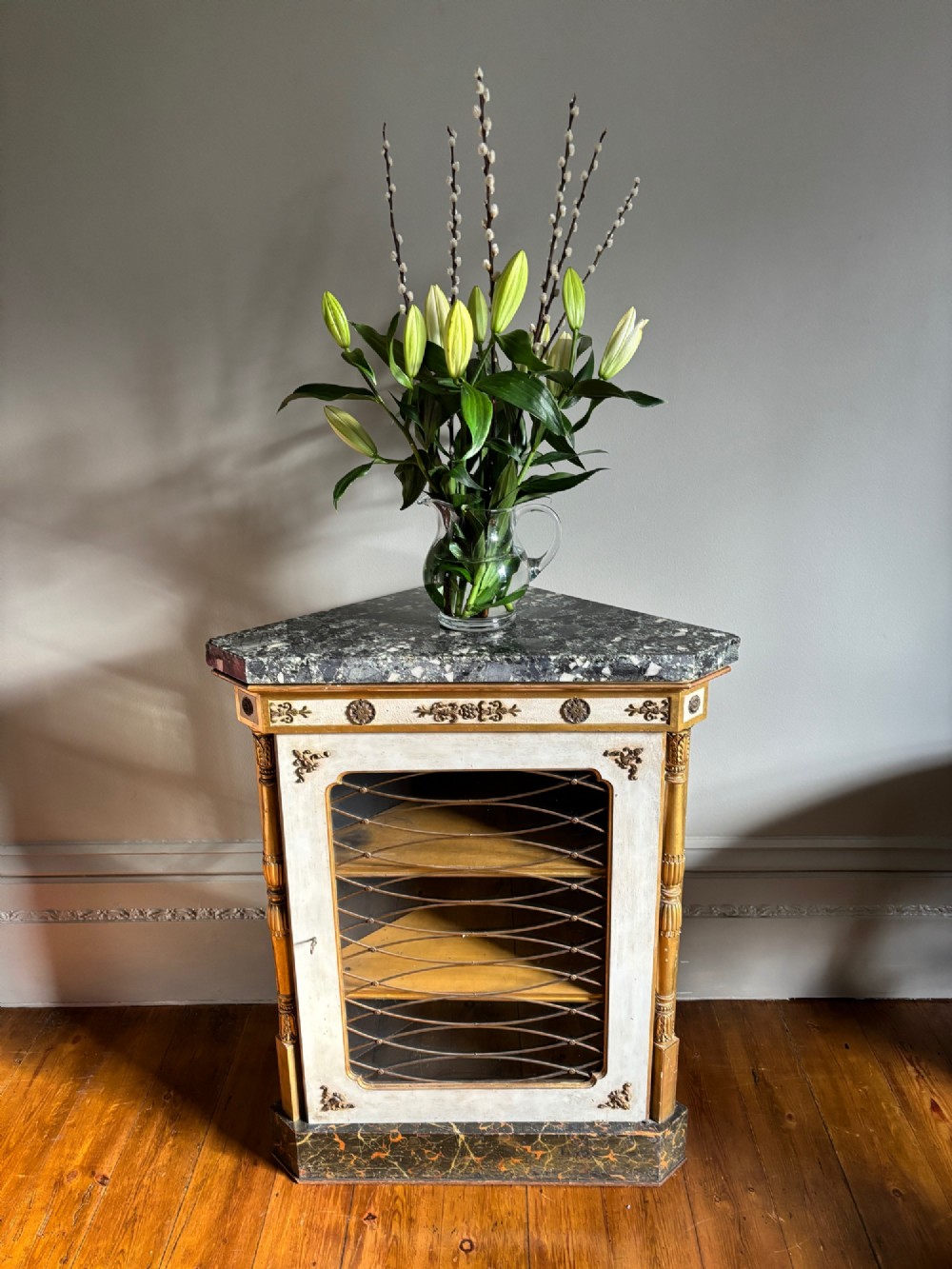 An Italian Regency period corner cupboard featuring a grey marble top, cream painted door with a gilt metal grille, fluted side columns, and decorative gilt rosettes. View VI