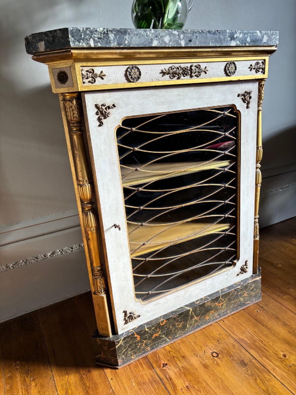An Italian Regency period corner cupboard featuring a grey marble top, cream painted door with a gilt metal grille, fluted side columns, and decorative gilt rosettes. View VIII
