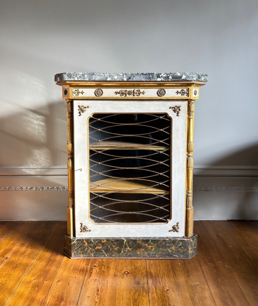 An Italian Regency period corner cupboard featuring a grey marble top, cream painted door with a gilt metal grille, fluted side columns, and decorative gilt rosettes. View IX