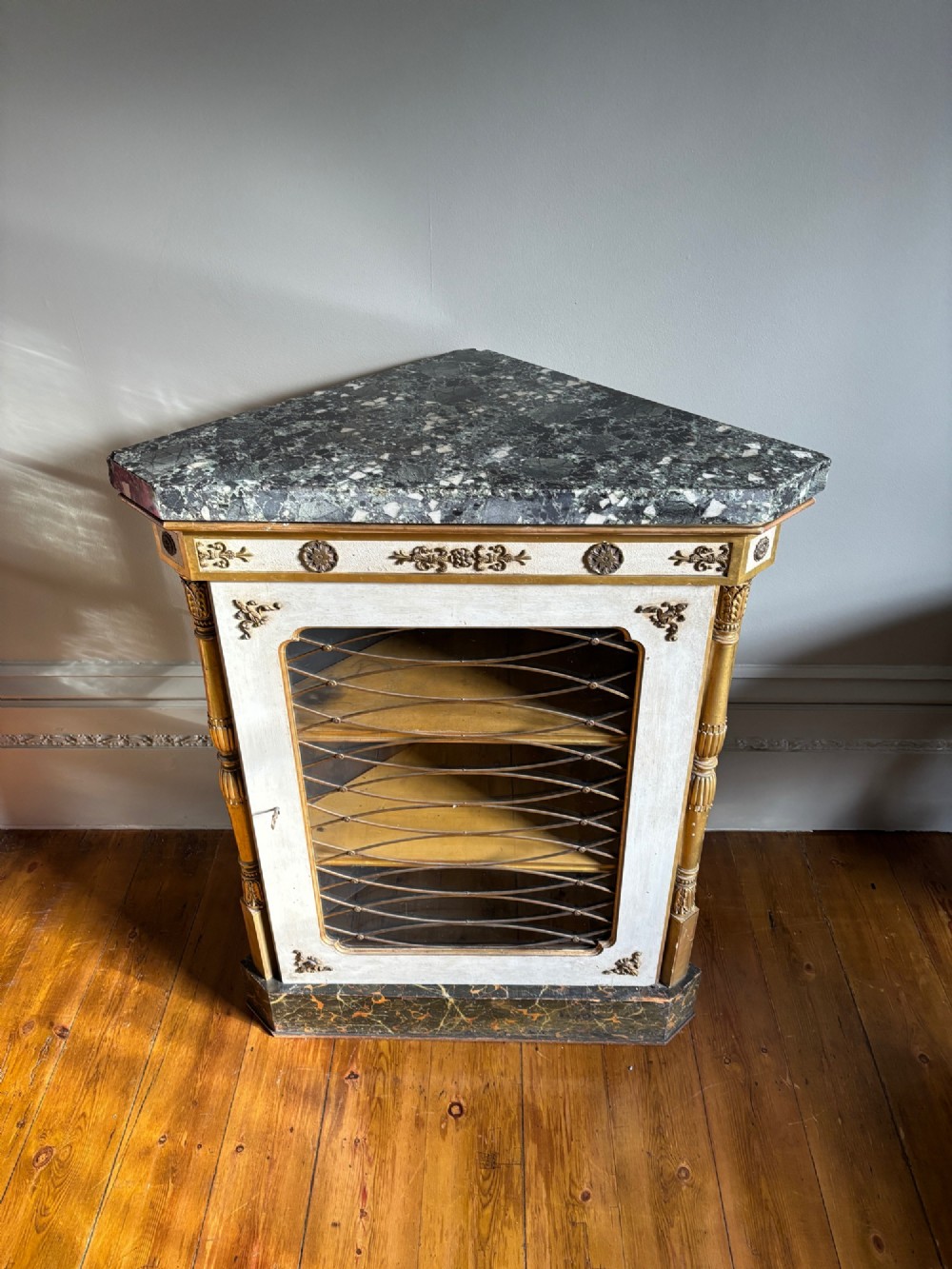 An Italian Regency period corner cupboard featuring a grey marble top, cream painted door with a gilt metal grille, fluted side columns, and decorative gilt rosettes. View X