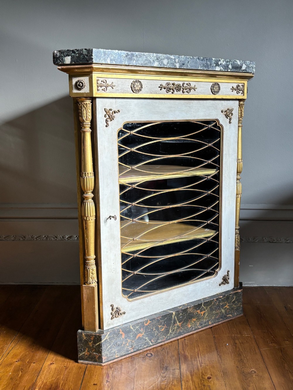 An Italian Regency period corner cupboard featuring a grey marble top, cream painted door with a gilt metal grille, fluted side columns, and decorative gilt rosettes.