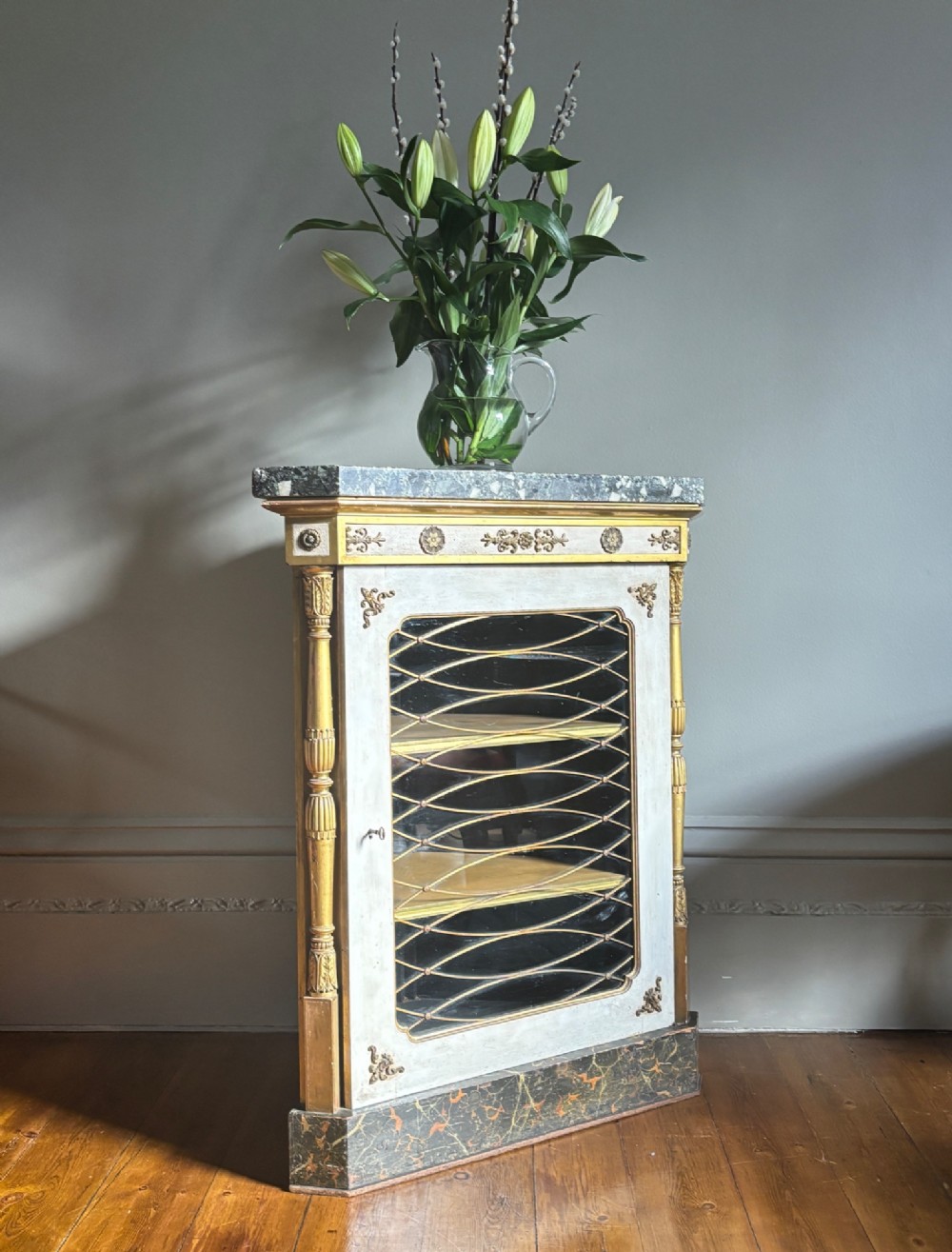 An Italian Regency period corner cupboard featuring a grey marble top, cream painted door with a gilt metal grille, fluted side columns, and decorative gilt rosettes. View II