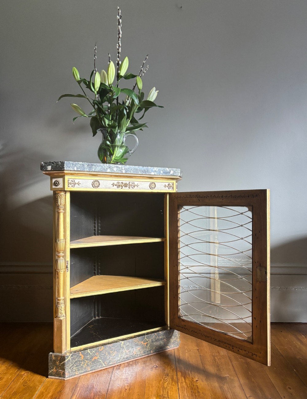 An Italian Regency period corner cupboard featuring a grey marble top, cream painted door with a gilt metal grille, fluted side columns, and decorative gilt rosettes. View XI