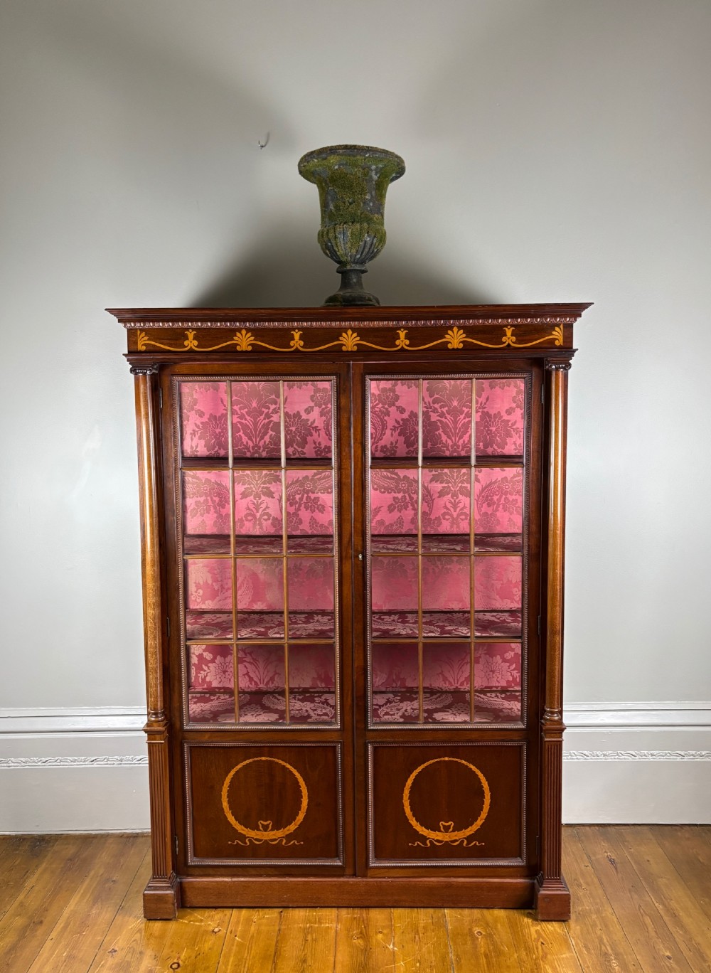 A tall dark rosewood display cabinet featuring two glazed doors with pink damask fabric lining, flanked by reeded side columns and decorated with inlaid frieze swags and wreath motifs. View II
