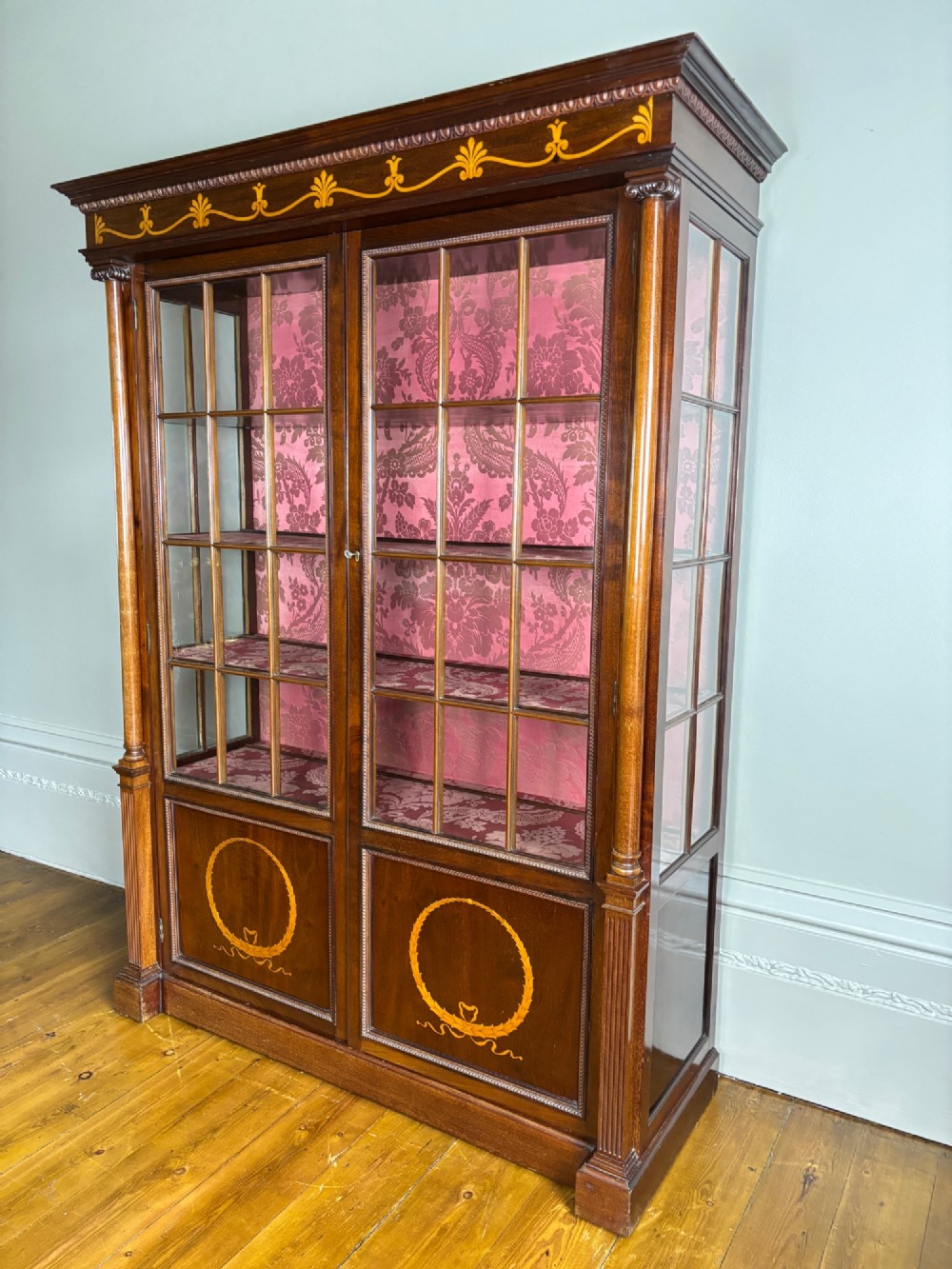 A tall dark rosewood display cabinet featuring two glazed doors with pink damask fabric lining, flanked by reeded side columns and decorated with inlaid frieze swags and wreath motifs. View III