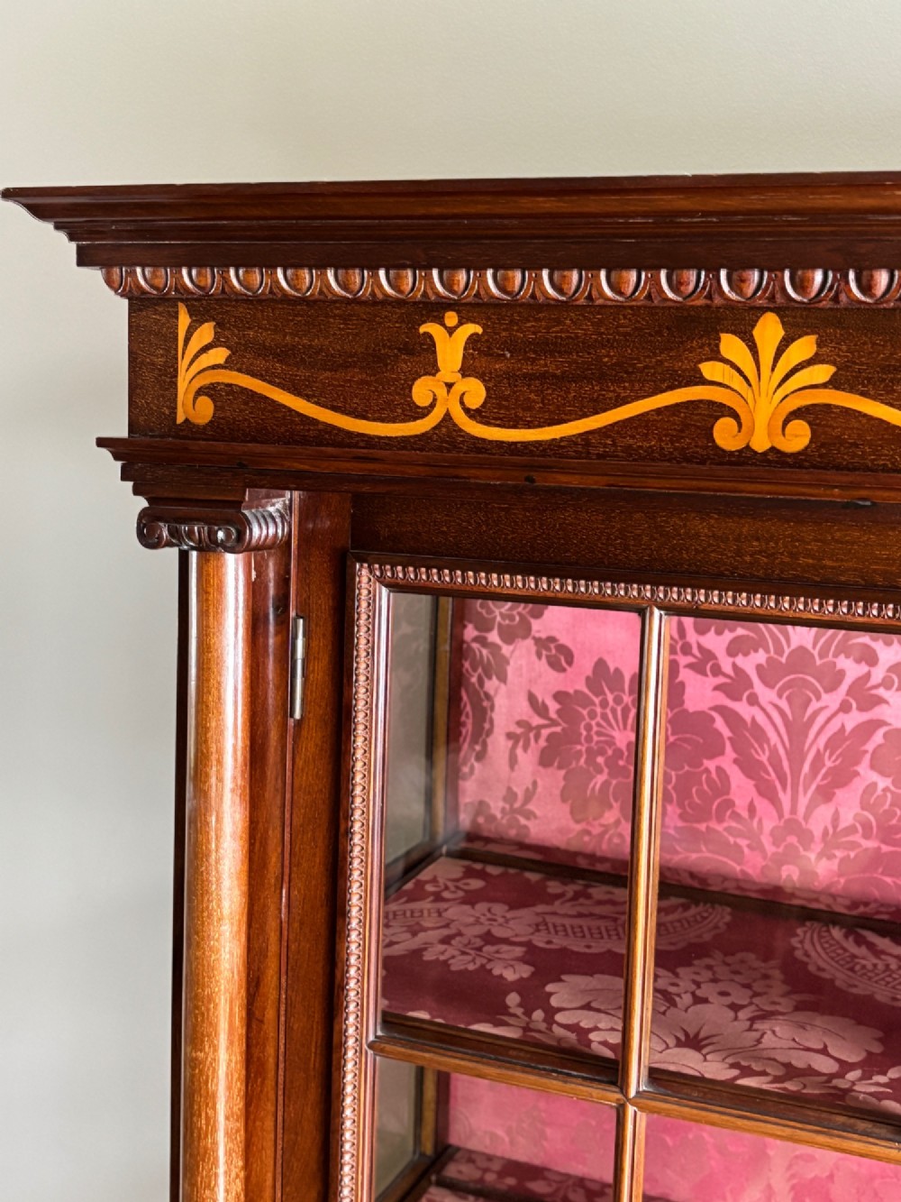 A tall dark rosewood display cabinet featuring two glazed doors with pink damask fabric lining, flanked by reeded side columns and decorated with inlaid frieze swags and wreath motifs. View V