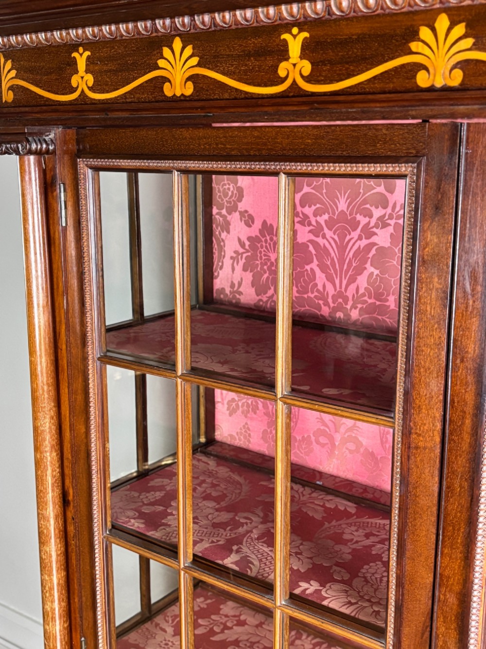 A tall dark rosewood display cabinet featuring two glazed doors with pink damask fabric lining, flanked by reeded side columns and decorated with inlaid frieze swags and wreath motifs. View VI