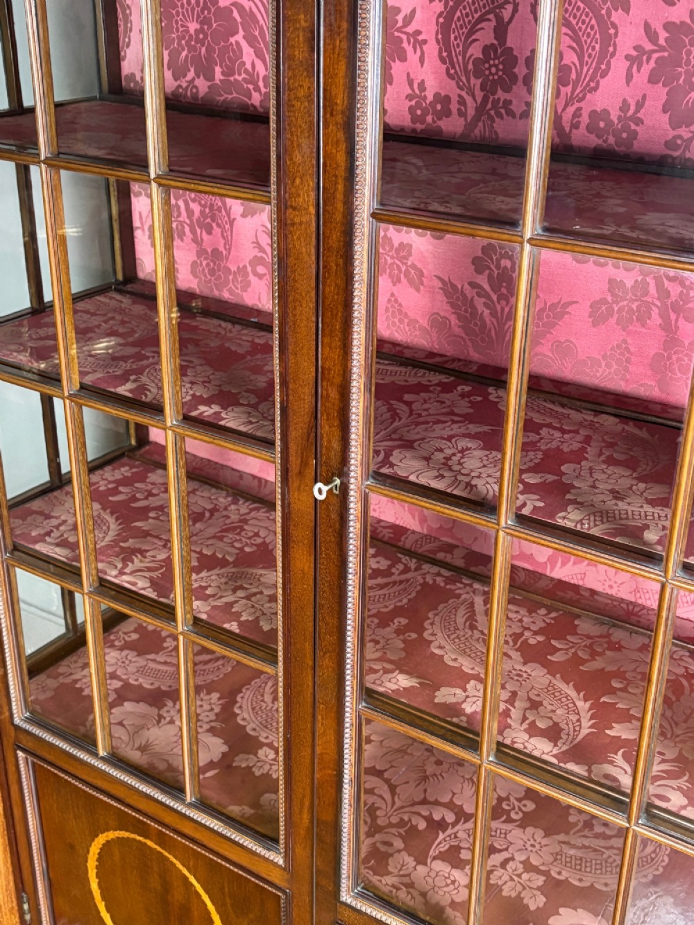 A tall dark rosewood display cabinet featuring two glazed doors with pink damask fabric lining, flanked by reeded side columns and decorated with inlaid frieze swags and wreath motifs. View VII