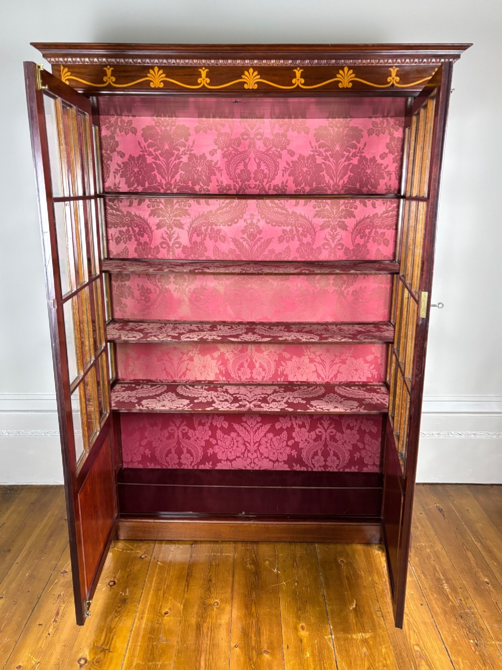 A tall dark rosewood display cabinet featuring two glazed doors with pink damask fabric lining, flanked by reeded side columns and decorated with inlaid frieze swags and wreath motifs. View IX