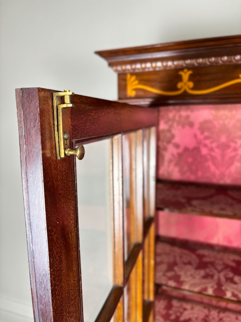 A tall dark rosewood display cabinet featuring two glazed doors with pink damask fabric lining, flanked by reeded side columns and decorated with inlaid frieze swags and wreath motifs. View X