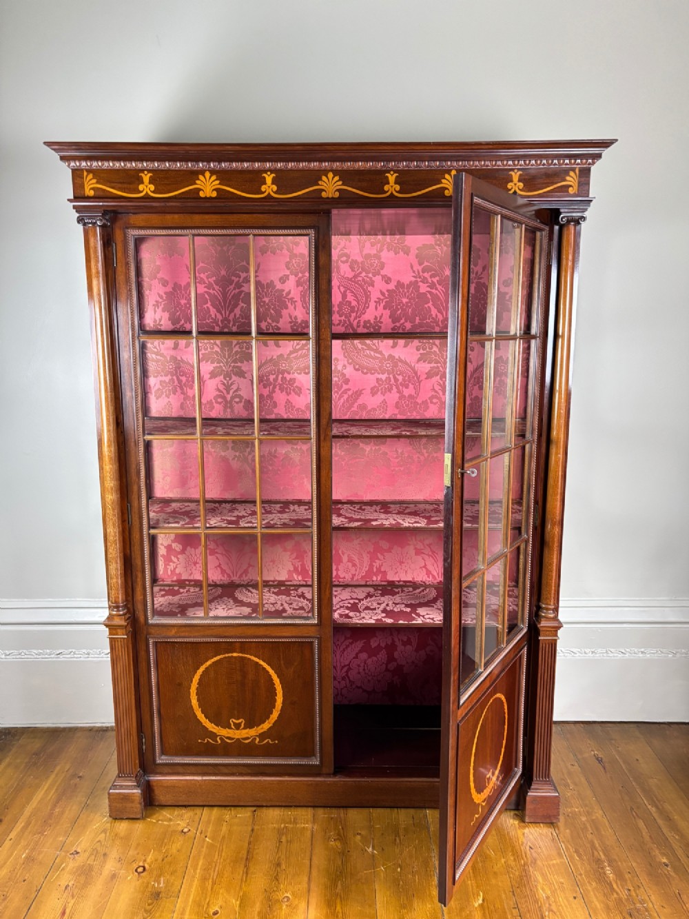 A tall dark rosewood display cabinet featuring two glazed doors with pink damask fabric lining, flanked by reeded side columns and decorated with inlaid frieze swags and wreath motifs. View XI