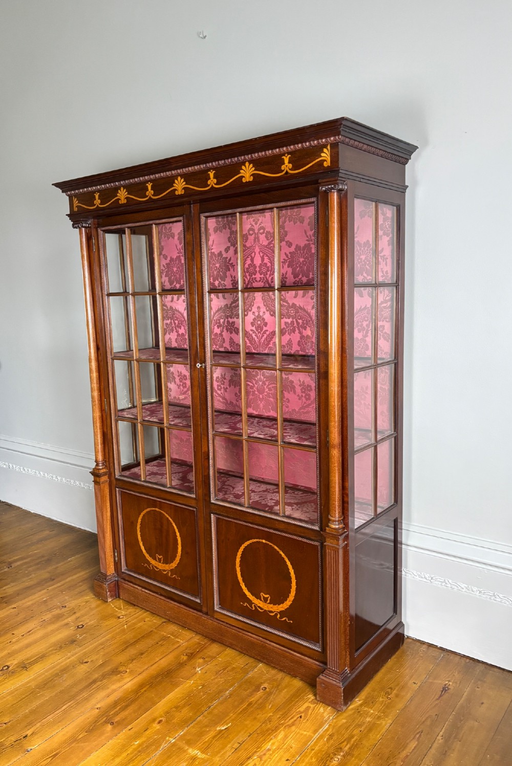 A tall dark rosewood display cabinet featuring two glazed doors with pink damask fabric lining, flanked by reeded side columns and decorated with inlaid frieze swags and wreath motifs. View XV