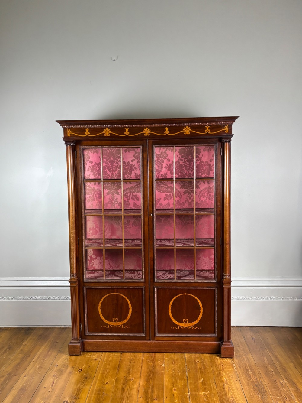 A tall dark rosewood display cabinet featuring two glazed doors with pink damask fabric lining, flanked by reeded side columns and decorated with inlaid frieze swags and wreath motifs.