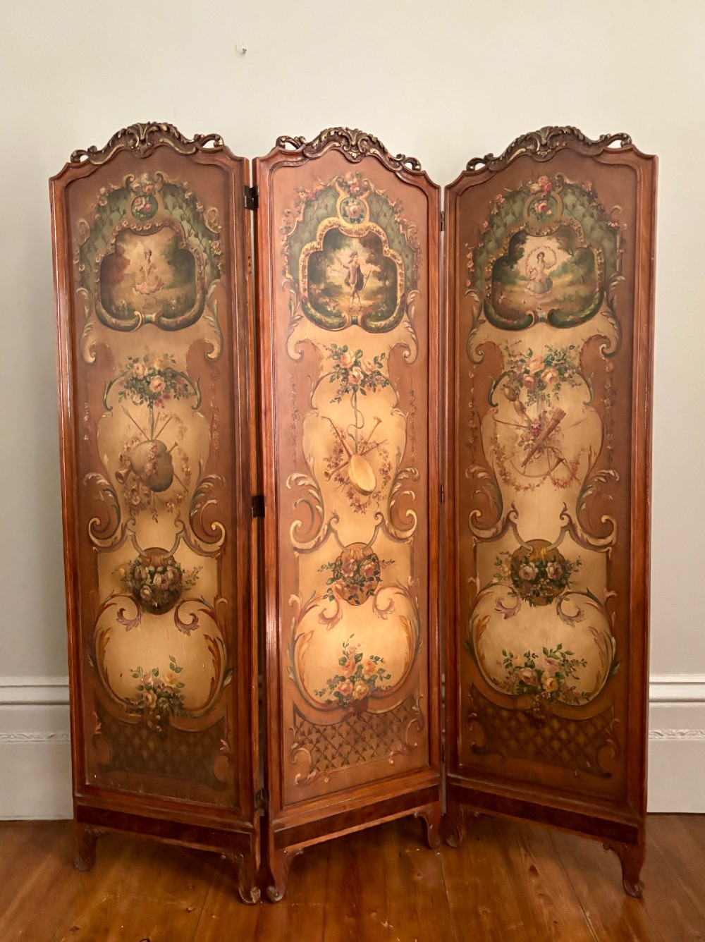 A French three-fold walnut screen featuring carved crestings and hand-painted panels depicting pastoral scenes, floral garlands, and musical trophies.