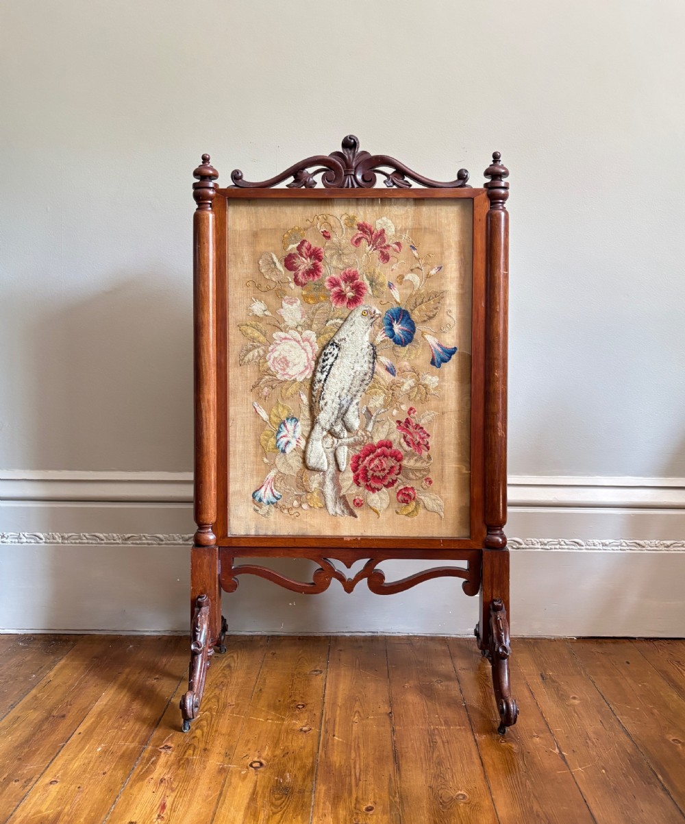 A Victorian dark timber fire screen with turned supports featuring a central embroidered panel of a bird amidst colourful flowers.