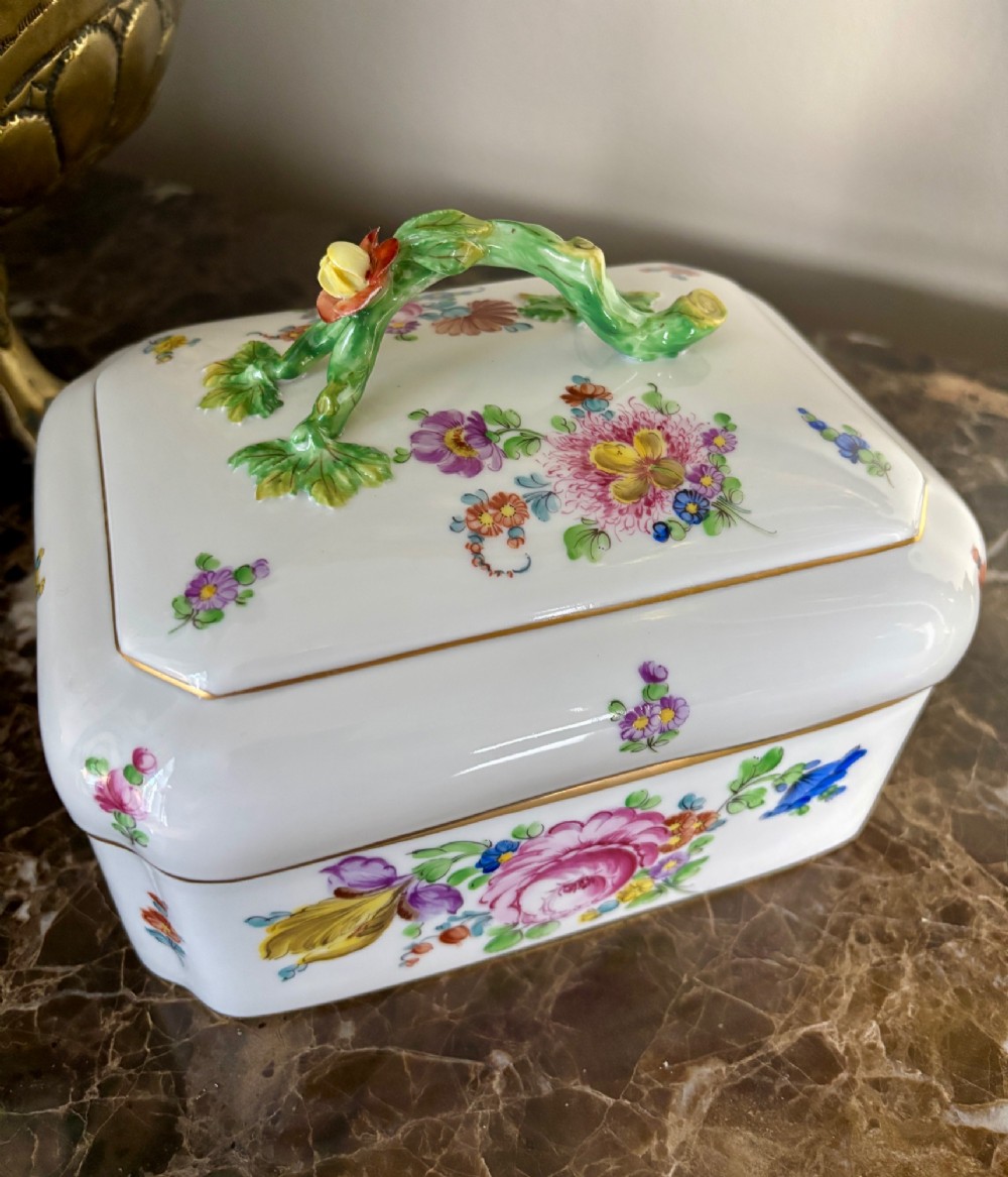 A rectangular white porcelain lidded box with canted corners, featuring hand-painted floral sprays, gilt rims, and a moulded green vine handle. View II