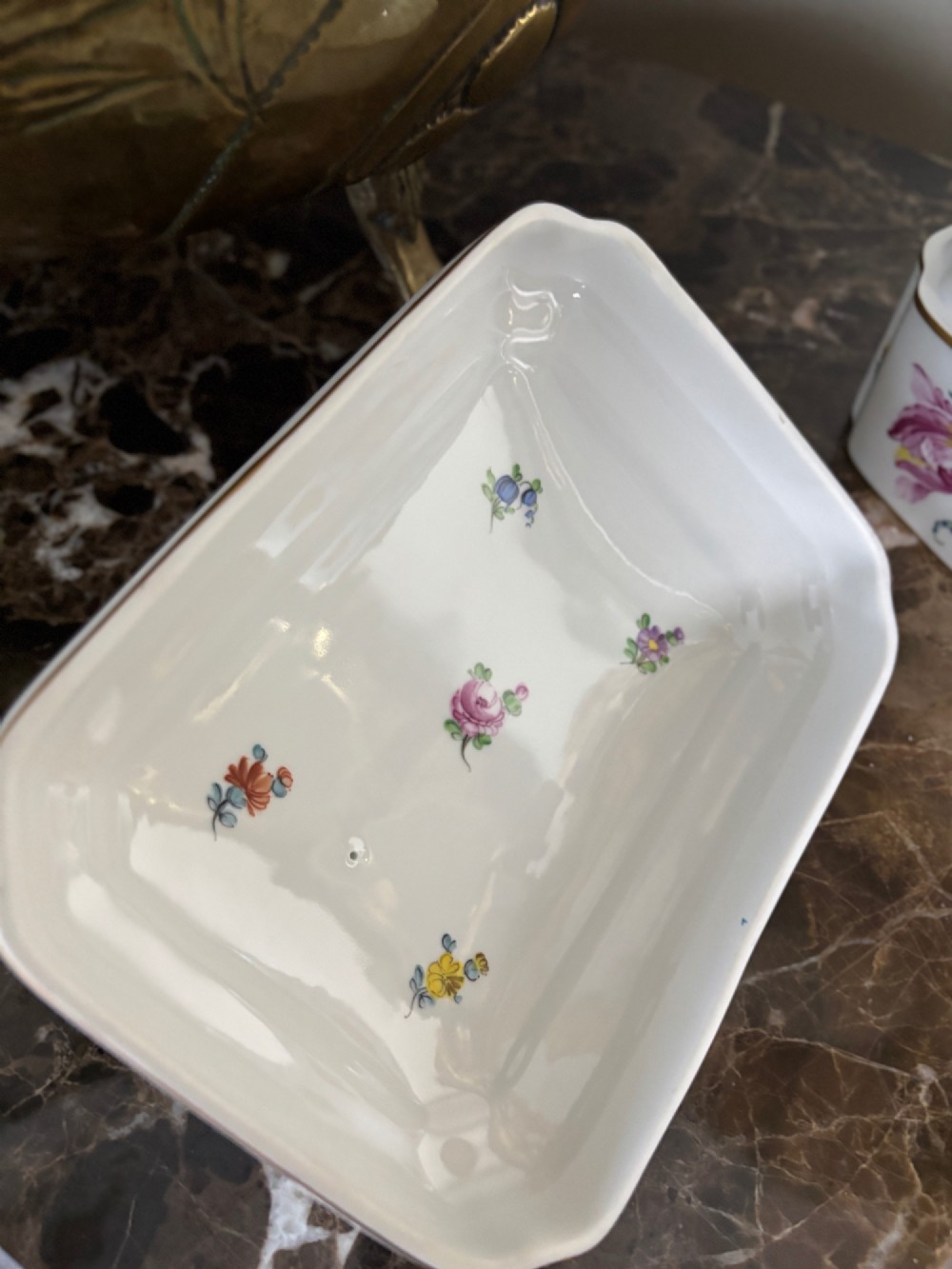 A rectangular white porcelain lidded box with canted corners, featuring hand-painted floral sprays, gilt rims, and a moulded green vine handle. View V