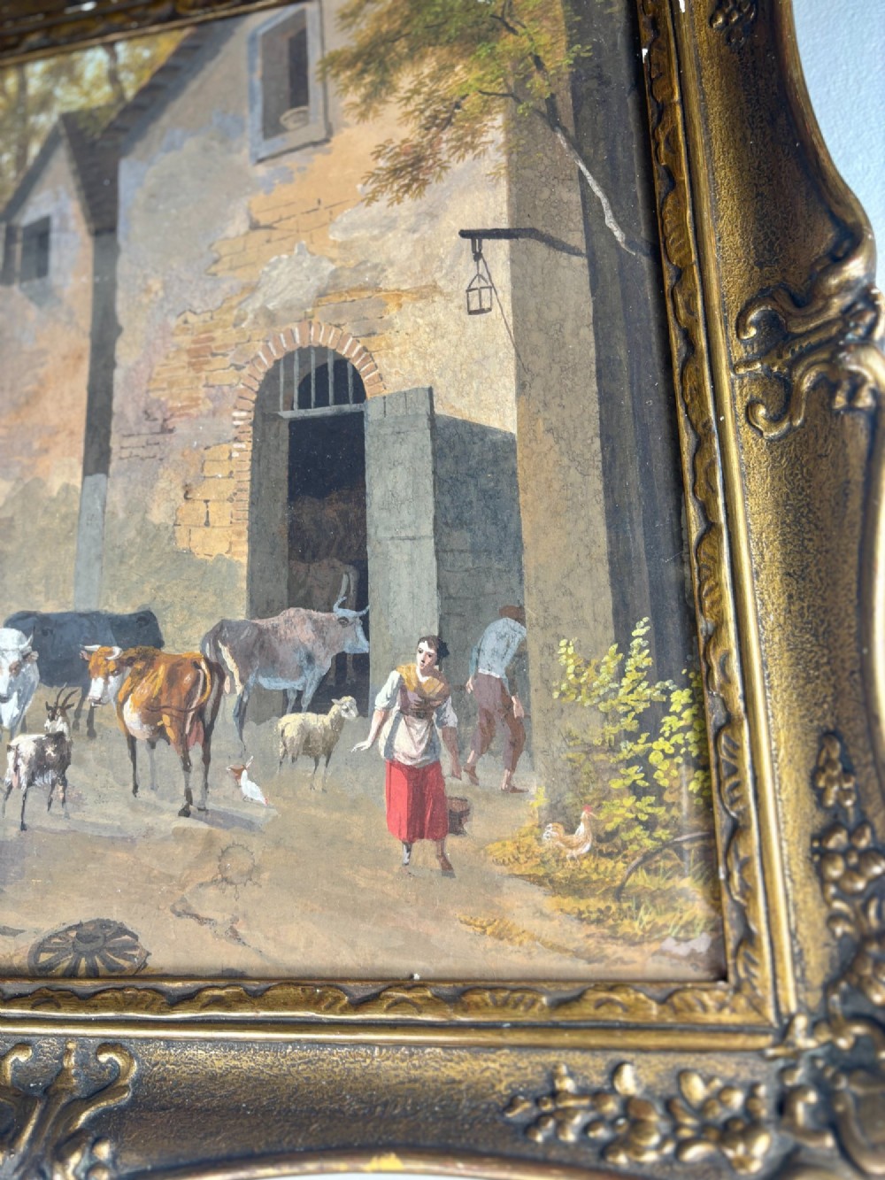 A framed gouache painting depicting a rural farmyard scene with livestock and rustic stone buildings set within an ornate gilded frame. View IV