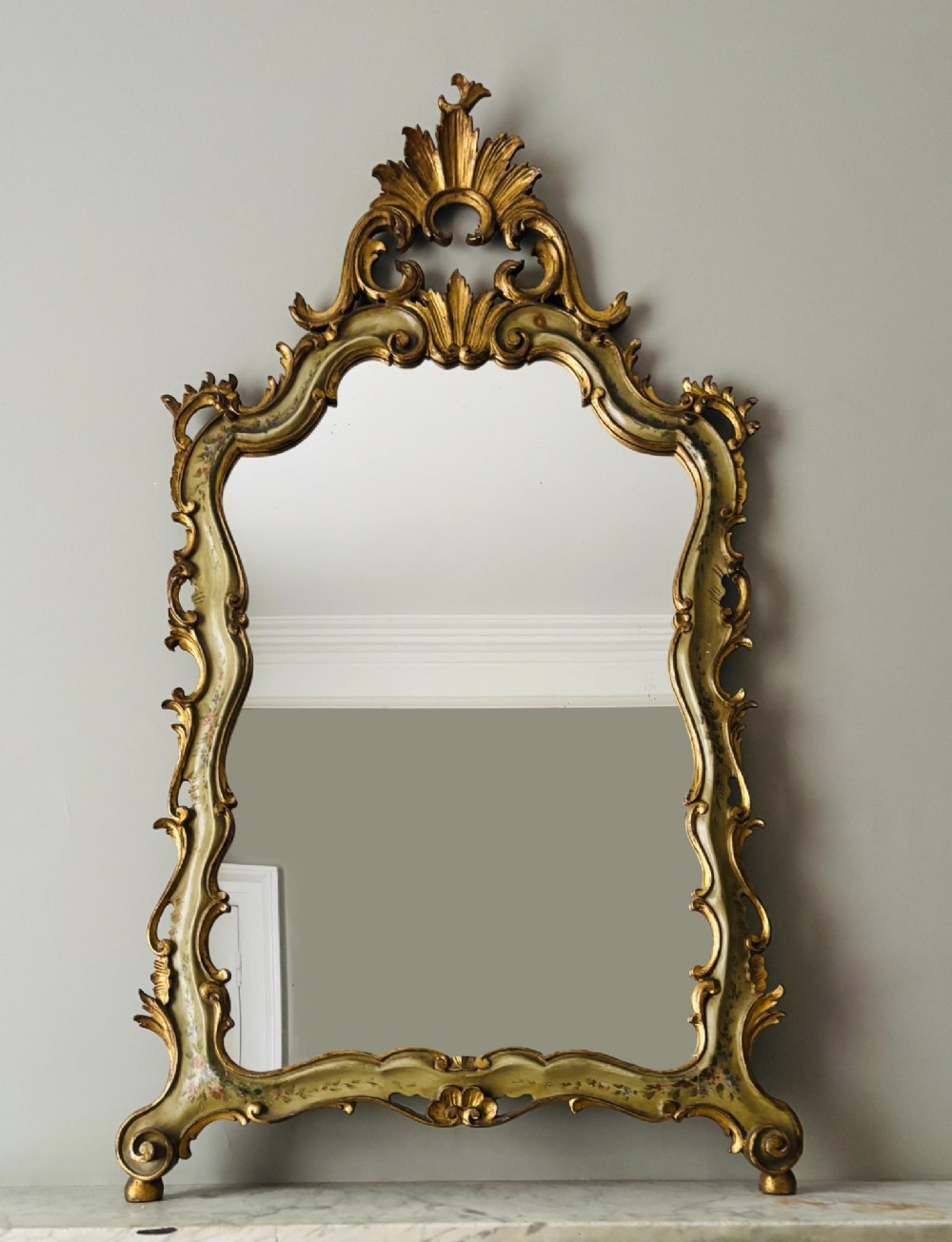 A mid-19th century Italian Venetian wall mirror featuring a carved giltwood frame with scrolling cresting, a sage green polychrome finish, and hand-painted floral detail.