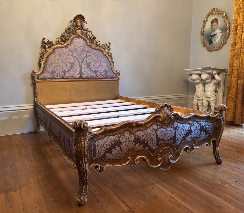An Italian 19th-century Rococo style giltwood bed with shell crest carvings and purple floral damask upholstery on the headboard and footboard.