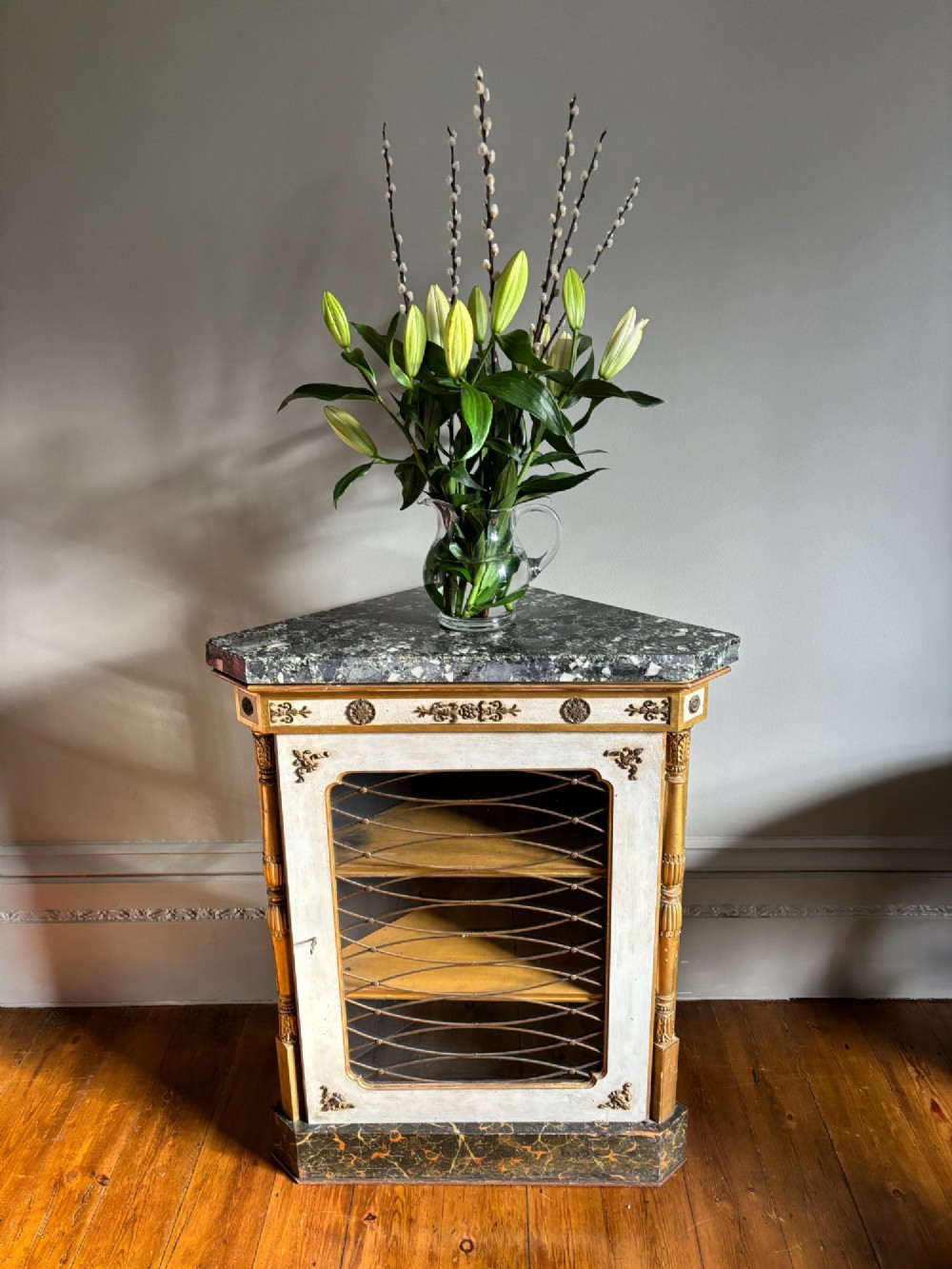 An Italian Regency period corner cupboard with a dark marble top, cream painted finish, gilded columns, and a single glazed door with decorative wire grille. View IV