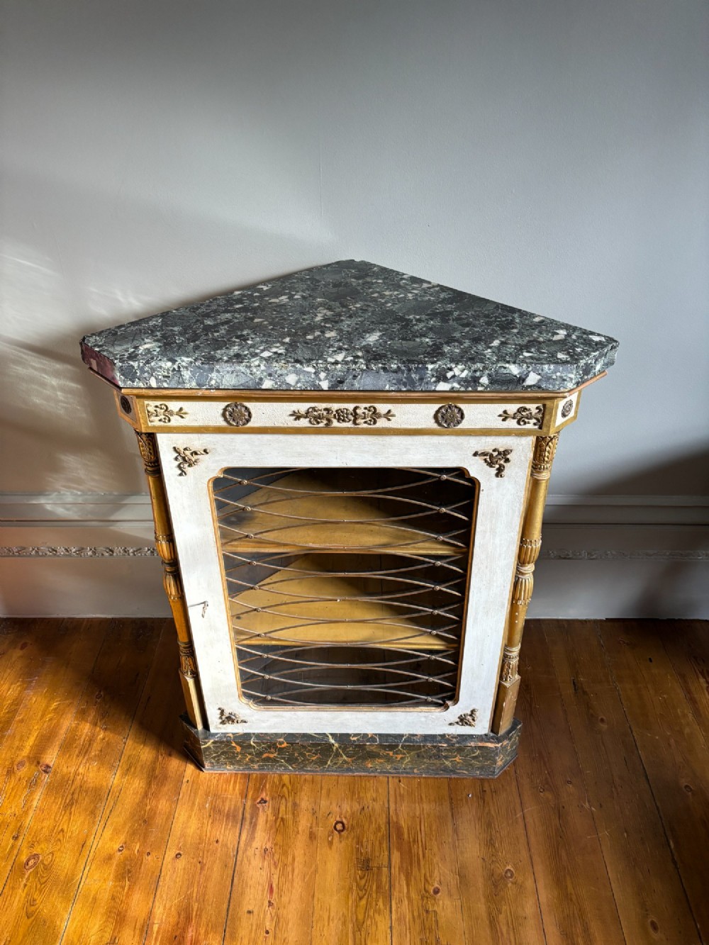 An Italian Regency period corner cupboard with a dark marble top, cream painted finish, gilded columns, and a single glazed door with decorative wire grille. View VIII