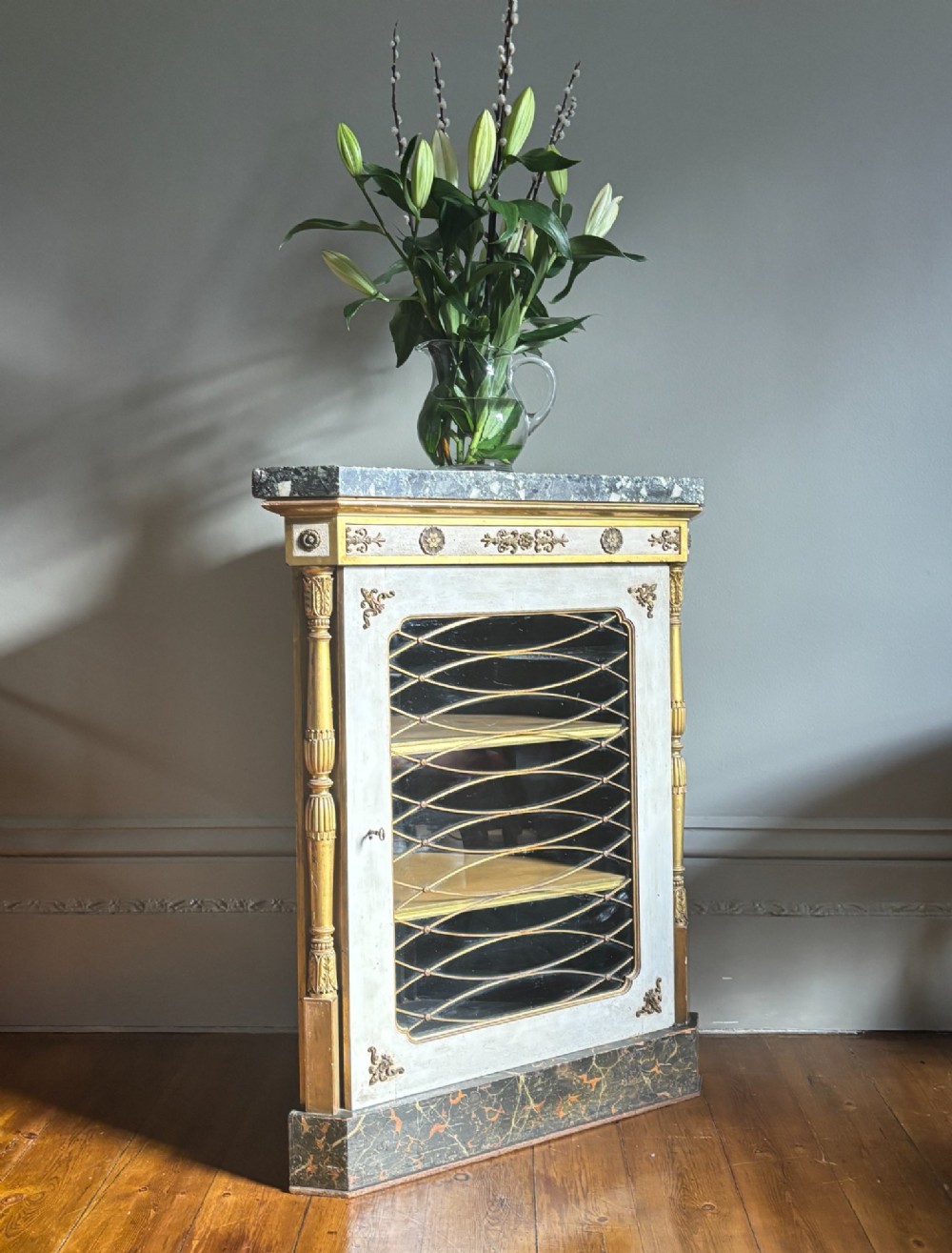 An Italian Regency period corner cupboard with a dark marble top, cream painted finish, gilded columns, and a single glazed door with decorative wire grille. View IX
