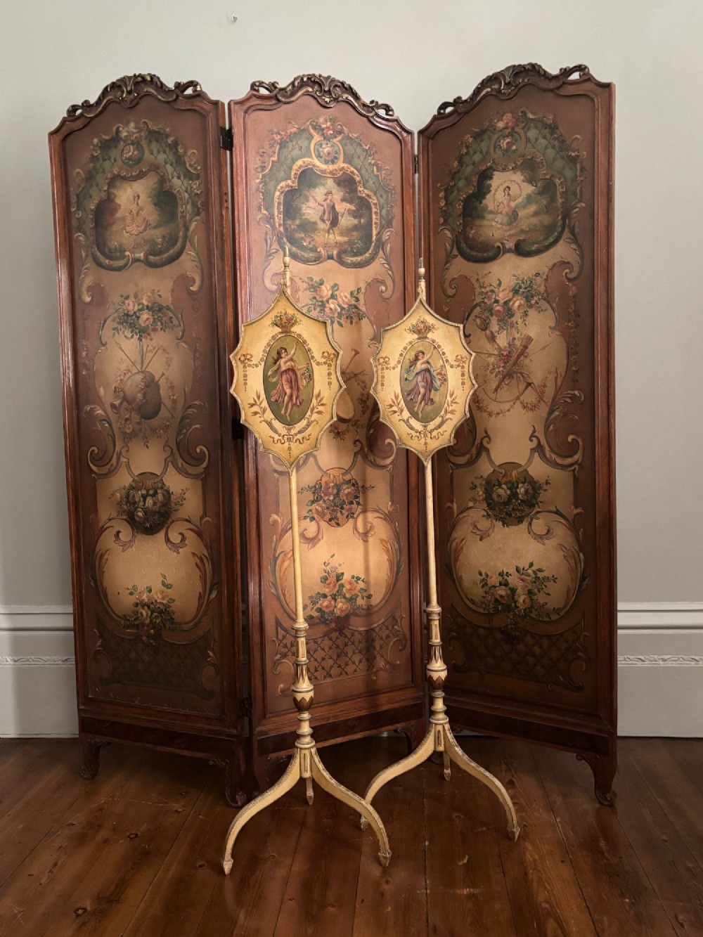 A pair of antique Georgian cream-painted wooden pole screens featuring hand-painted classical figures on shaped panels supported by tripod bases with spade feet. View II