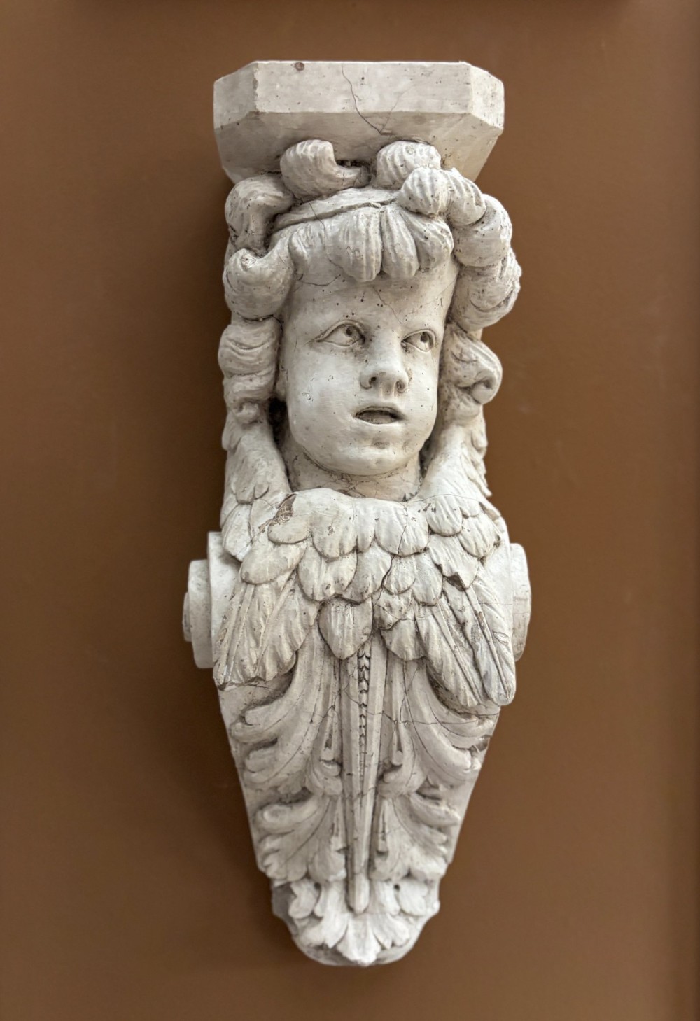 A carved off-white plaster wall corbel featuring a winged putto head above an acanthus leaf support.