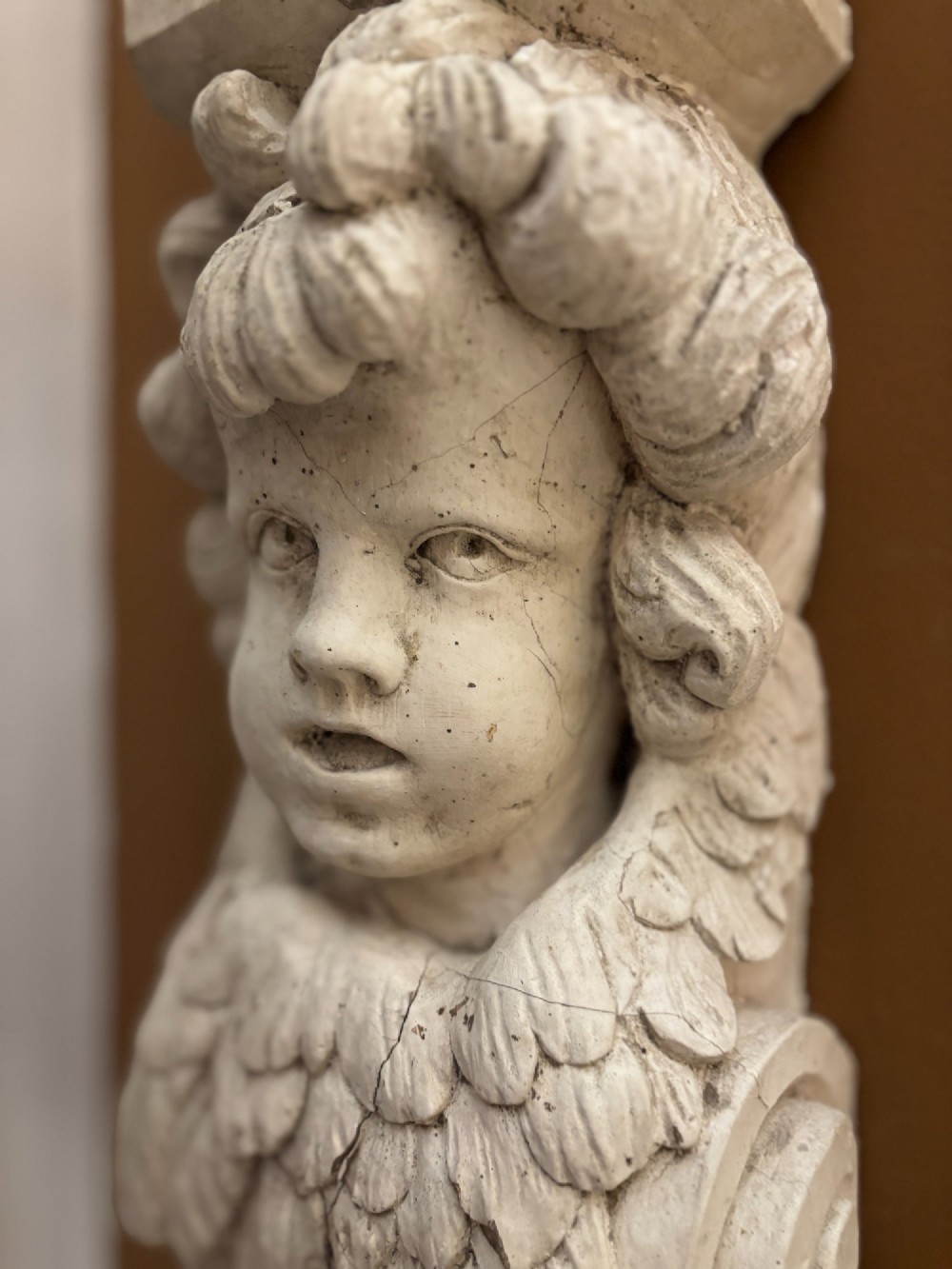 A carved off-white plaster wall corbel featuring a winged putto head above an acanthus leaf support. View III