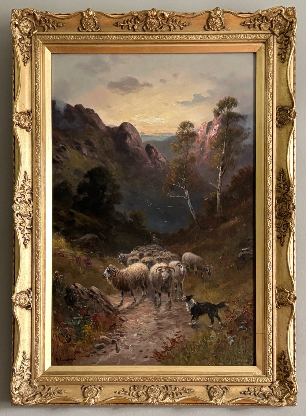 A vertical oil painting depicting a flock of sheep and a dog walking through a mountainous valley path at sunset, framed in an ornate gilded frame. View II