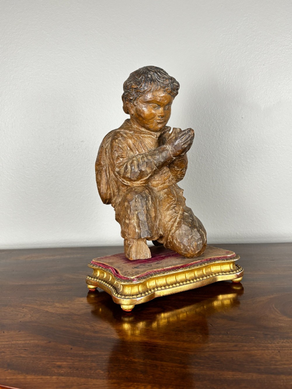 A hand-carved lime wood figure of a kneeling child in a praying pose, set on an ornate gilded wooden plinth with a red velvet top.