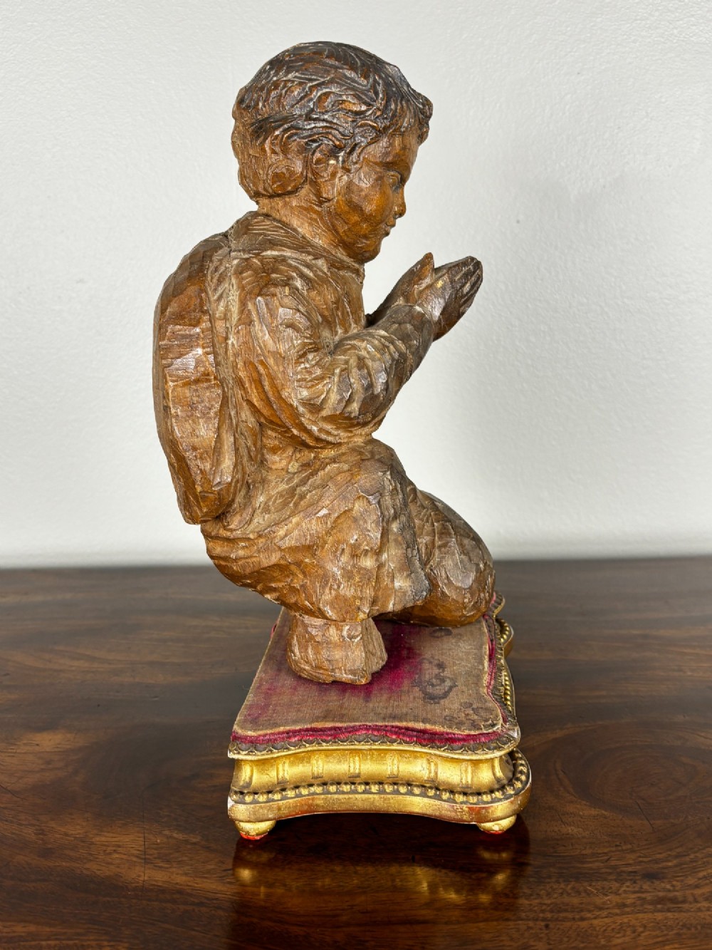 A hand-carved lime wood figure of a kneeling child in a praying pose, set on an ornate gilded wooden plinth with a red velvet top. View VI