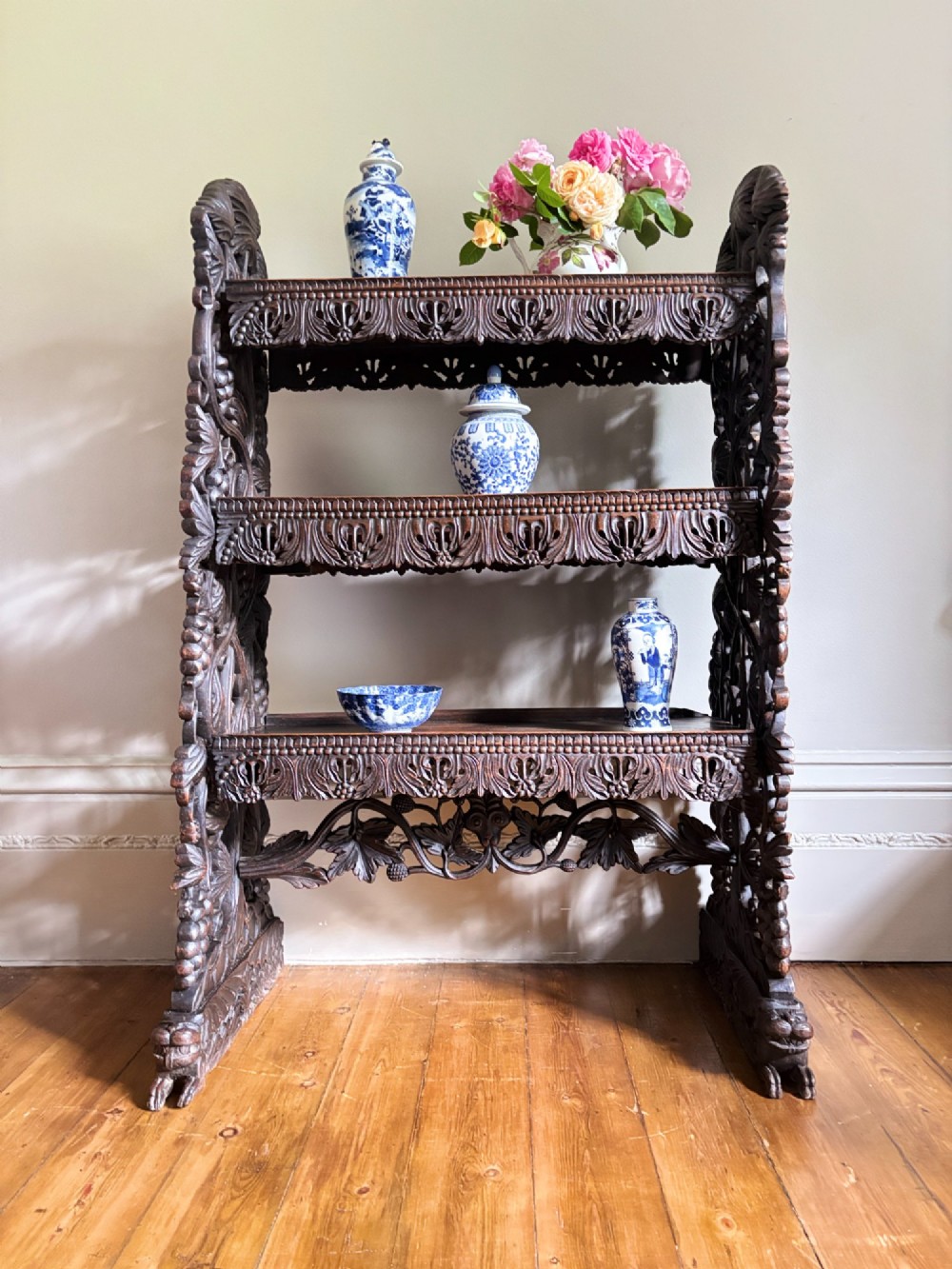 A mid-19th-century Anglo-Indian dark padauk wood open shelving unit with three tiers, intricate foliate carving, pierced aprons, and carved feet.