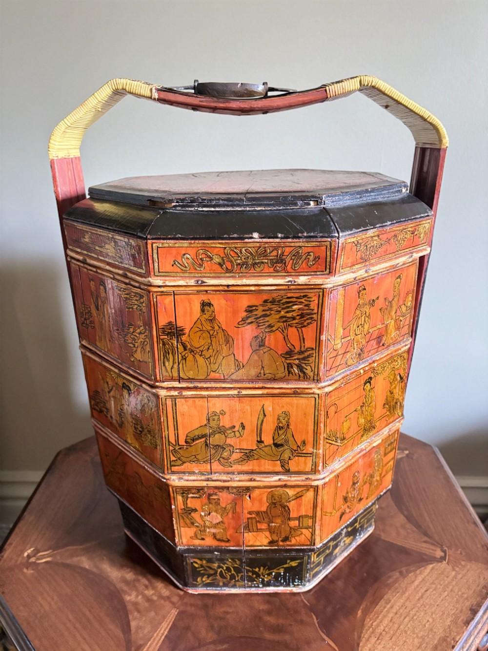 An octagonal three-tiered Chinese lacquer dowry basket decorated with hand-painted narrative panels and a high arched handle. View II