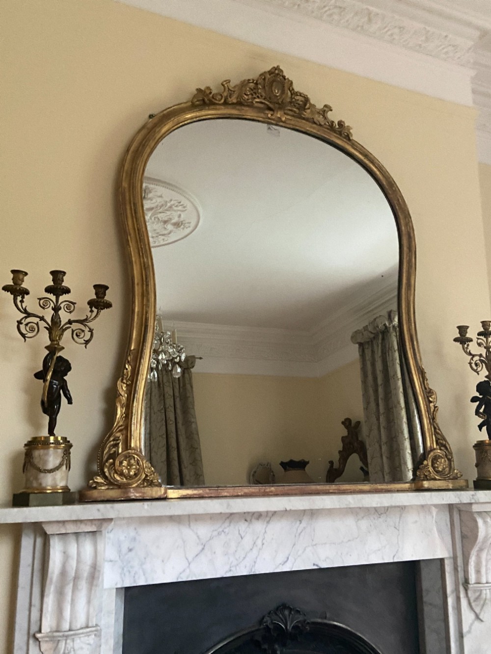 A large mid-nineteenth century giltwood overmantel mirror featuring a waisted arched frame, carved pediment cresting, and scroll motifs at the base. View V