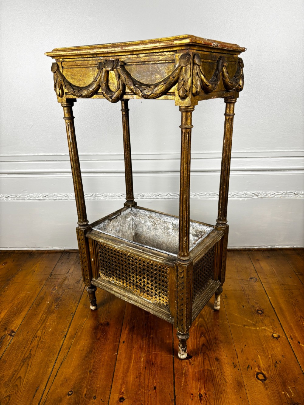 A two-tiered French Empire giltwood jardinere stand featuring carved festoons, fluted supports, and a lower caned trough with a removable metal liner.