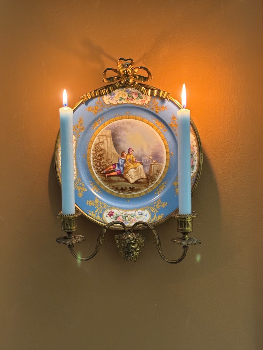 A 19th-century French wall candelabra featuring a sky blue porcelain plate with a central hand-painted scene, mounted in gilt ormolu with a top ribbon bow, a lower mask, and two curved candle arms holding lighted candles.