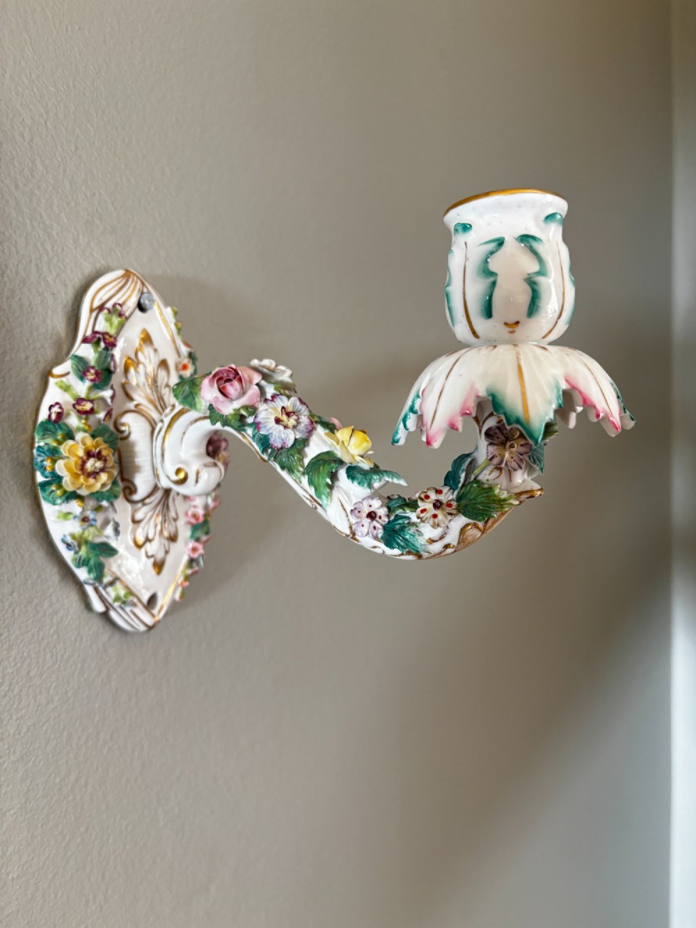 A white porcelain single candle wall sconce with hand-painted applied floral decorations, green foliage, and gilded Rococo scrollwork.