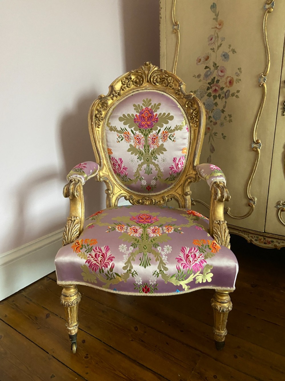 A carved giltwood open armchair upholstered in pink floral brocade silk featuring turned legs on castors.