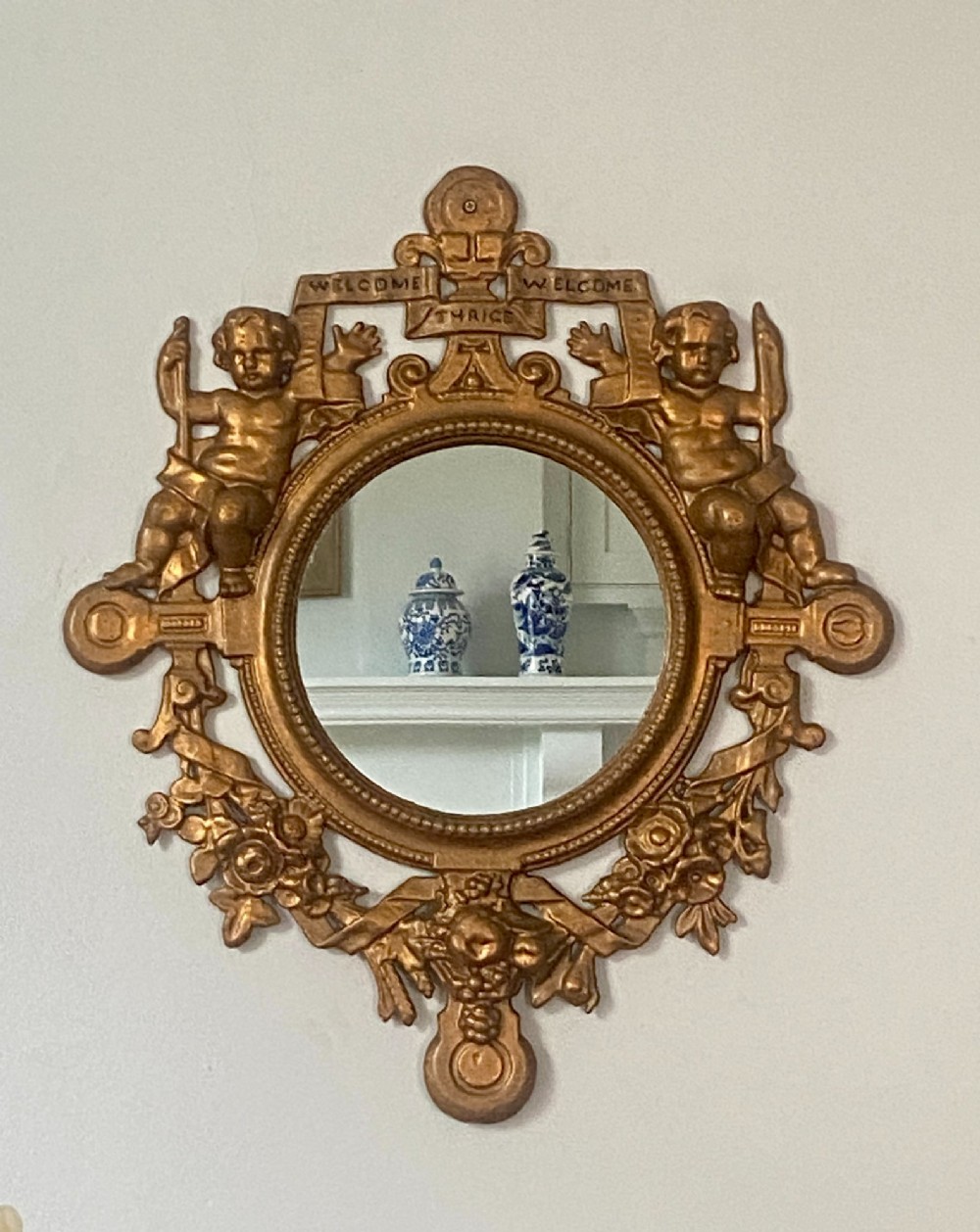 A circular gold-painted cast iron wall mirror with cherub figures, floral garlands, and a banner reading Welcome Thrice Welcome.