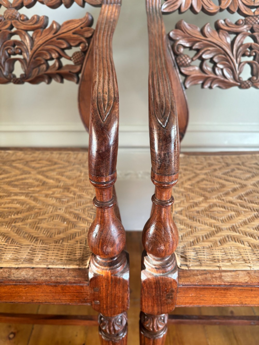 A pair of carved dark wood armchairs featuring openwork foliage backrests, curved arms, turned front legs, and woven cane seats. View VI