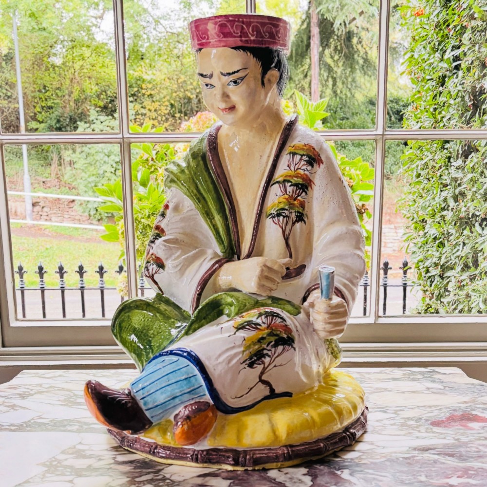 A polychrome glazed Italian ceramic figure of a seated Chinese scholar dressed in robes on a yellow cushion.