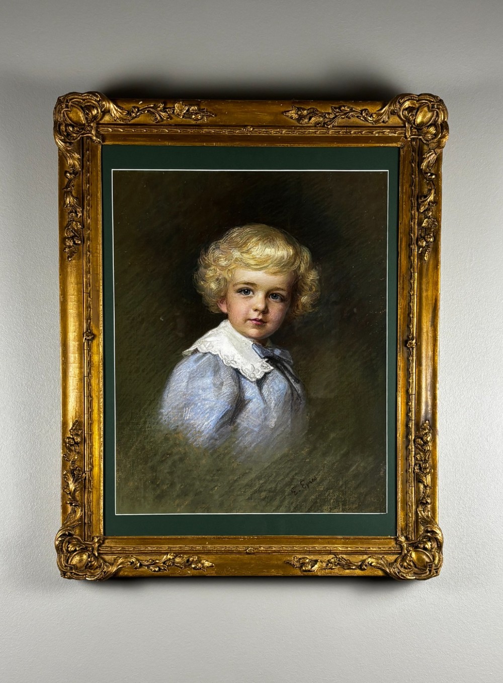 A framed pastel portrait of a young boy with blond curls wearing a light blue smock with a white lace collar, presented in an ornate gilt wood frame.