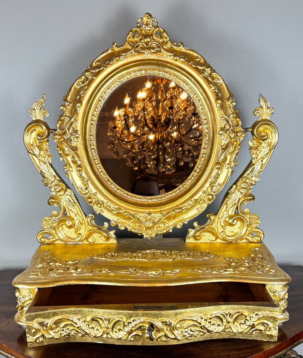 An 18th-century Italian Rococo giltwood and gesso table mirror with an oval glass frame supported by carved scrolling uprights above a base drawer.