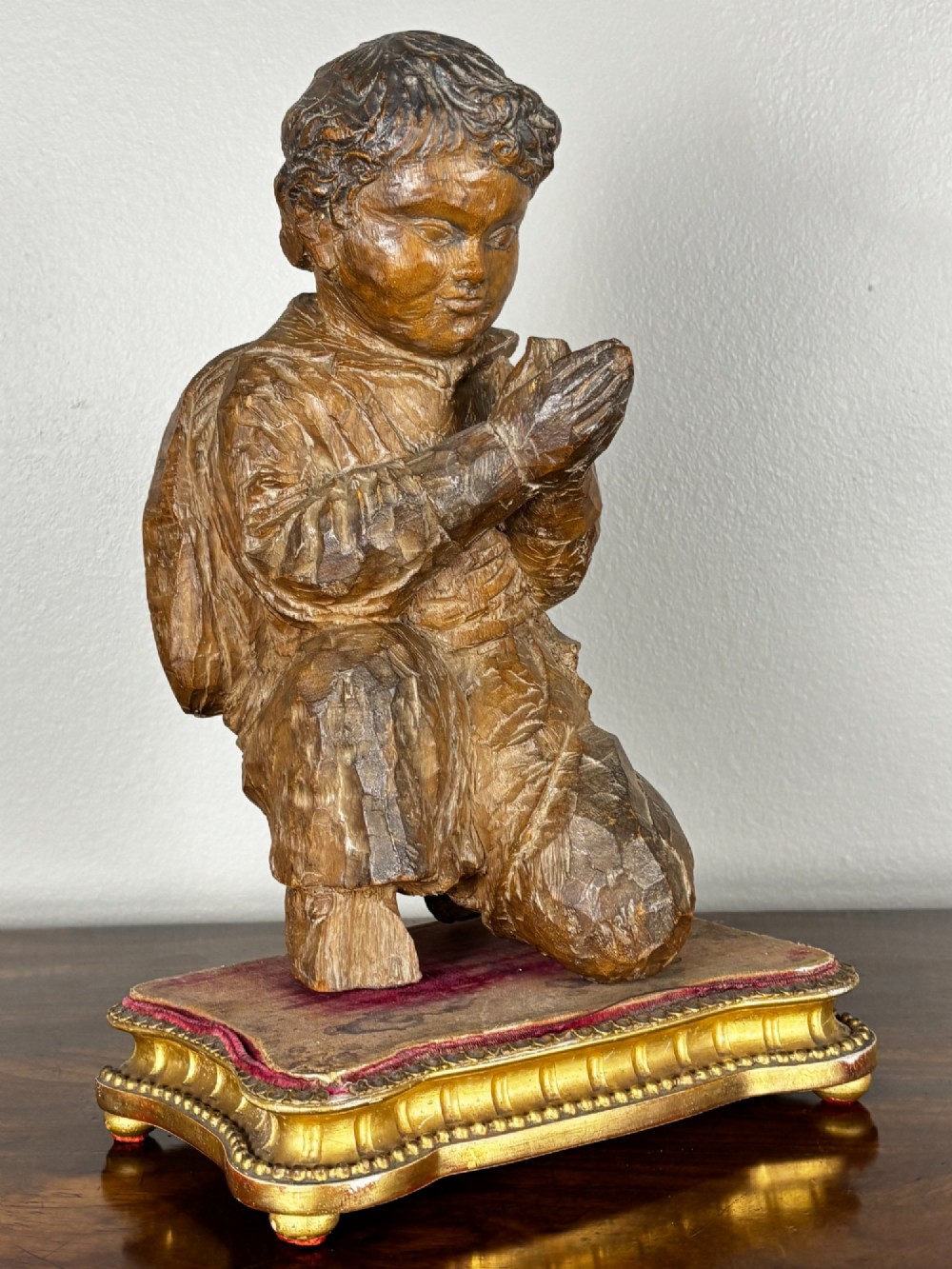 A late 18th-century carved limewood figure of a kneeling youth with hands folded in prayer, mounted on a velvet and gilded wooden base.
