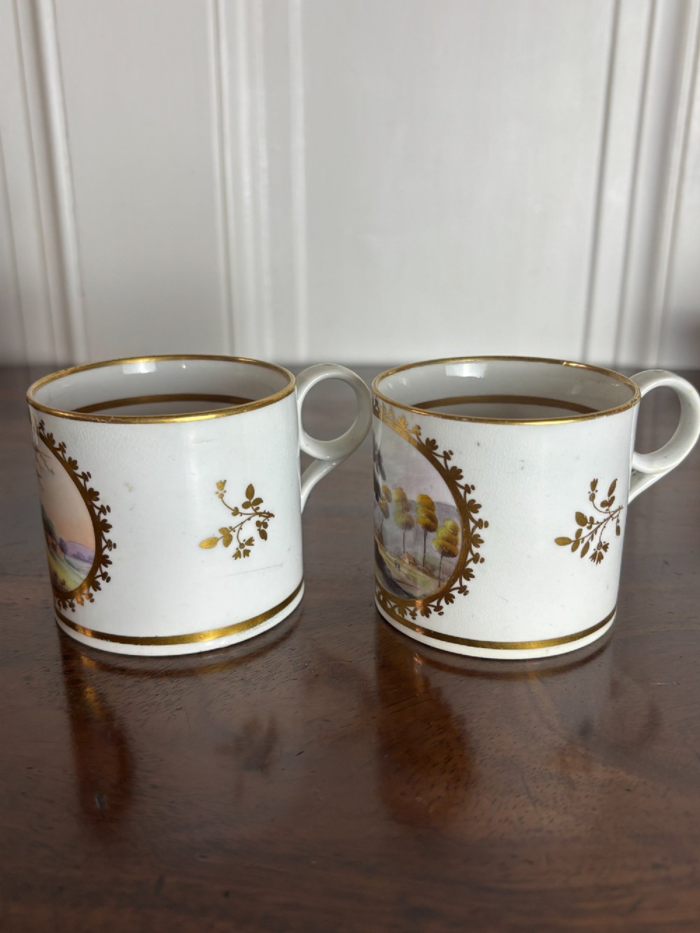 A pair of cylindrical white porcelain coffee cans decorated with hand-painted landscape vignettes enclosed in ornate gilded oval frames. View IV